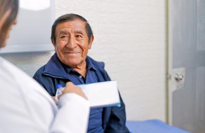 The image shows a person sitting indoors, wearing a dark blue collared shirt and a zip-up jacket. In the foreground, another individual dressed in a white coat is holding a clipboard or notepad and appears to be writing or taking notes. The setting looks like a clinical or consultation room, with a light-colored wall and a closed door in the background.