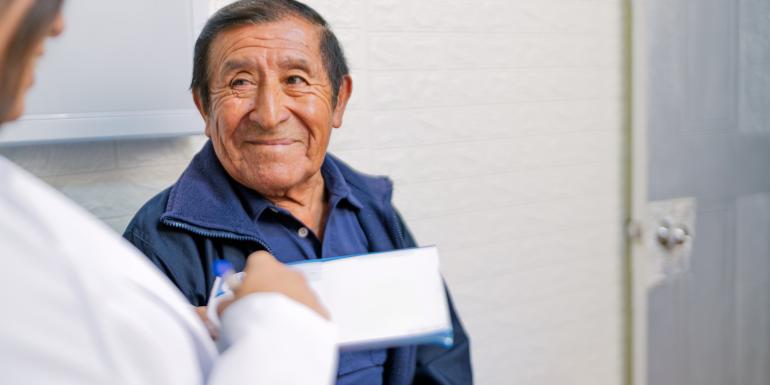 La imagen muestra a una persona sentada en una habitación interior, vestida con una camisa tipo polo azul oscuro y una chaqueta del mismo tono. En primer plano, se observa a otra persona con bata blanca sosteniendo una libreta o portapapeles, aparentemente tomando notas. El entorno parece ser una sala de consulta médica, con una pared clara y una puerta cerrada al fondo.