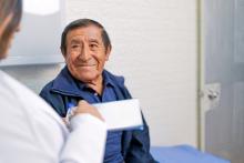 The image shows a person sitting indoors, wearing a dark blue collared shirt and a zip-up jacket. In the foreground, another individual dressed in a white coat is holding a clipboard or notepad and appears to be writing or taking notes. The setting looks like a clinical or consultation room, with a light-colored wall and a closed door in the background.